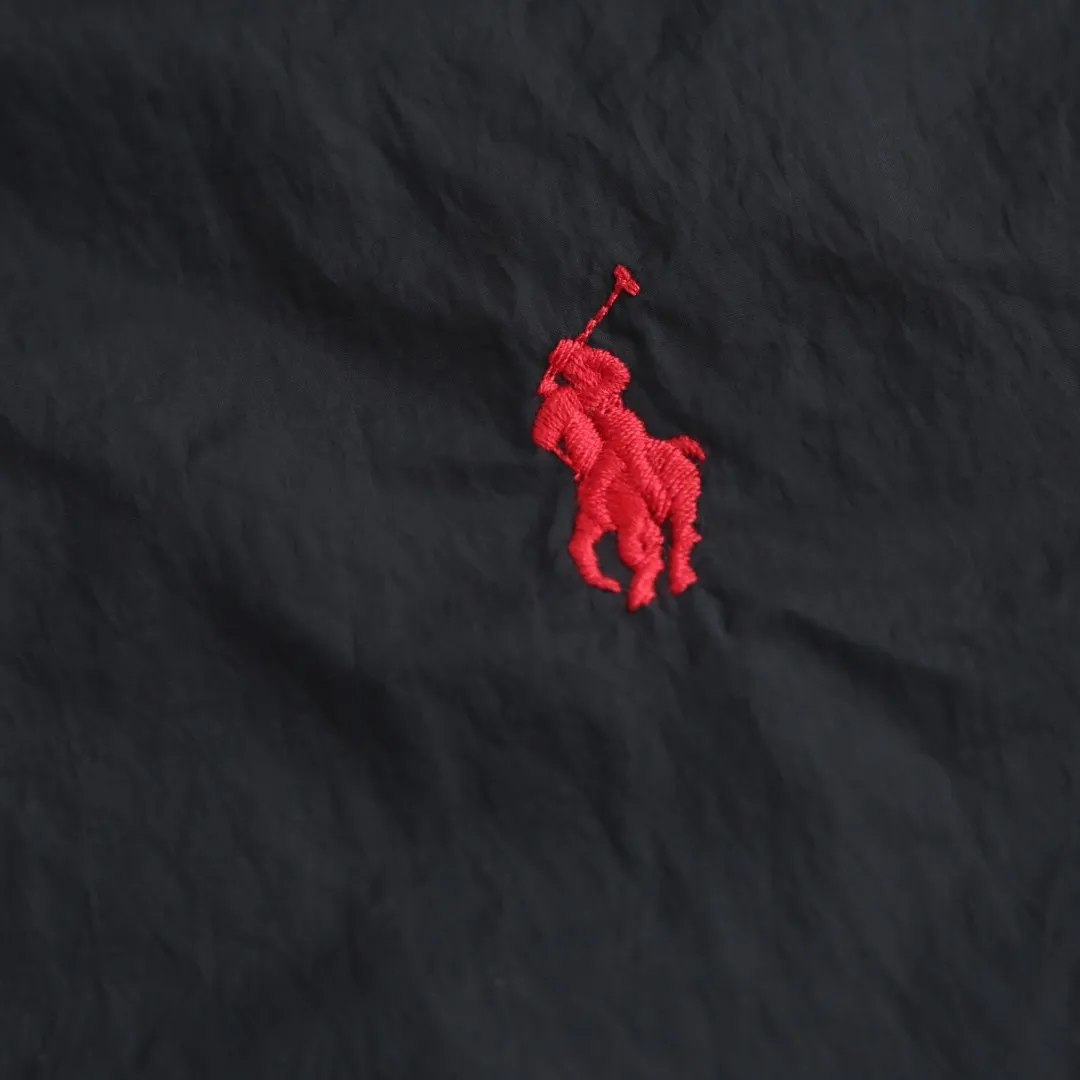 🅿️680 Ralph Lauren pony embroidered light cotton jacket. The fabric is made of high-density nylon taffeta fabric, which combines pragmatism and classic texture. The density is about 210T-300T, the surface is smooth and slightly matte, it has excellent windproof and waterproof performance (with DWR water-repellent treatment), it has strong wrinkle resistance and is easy to care for, which is in line with the positioning of American casual "durable and practical". Classic dark black, using vat dyeing process (dye penetrates into the fiber), the color fastness reaches ISO level 4 or above, preventing fading or color transfer after washing. The lining is made of light cotton-polyester blended fabric (65% Polyester + 35% Cotton) to reduce the friction between the fabric and the inner clothing and improve the smoothness of wearing; the blended material has both breathability and durability to avoid the stuffy feeling of pure nylon lining. The style is a loose fit of the classic Coach Jacket, with slightly dropped shoulder lines (shoulder width is 1-2cm wider than the standard fit). The length reaches the hips, and the sleeves are slightly tapered straight sleeves, taking into account both freedom of movement and casual style. The Polo horse logo on the left chest is flat satin stitch embroidered (satin embroidery), embroidery thread: 100% polyester high color fastness embroidery thread (the color number is consistent with the brand’s standard red color card), the stitch density is 12-15 stitches/cm, the outline of the horse logo is clear, and the details of the rider and horse are burr-free. The embroidery is positioned accurately through the template (12cm from the bottom of the neckline and 8cm from the placket) to ensure left-right symmetry and centering. The seams of the body are made of double-needle flat seams (needle pitch 3cm/10 needles), the stitches are flat and straight, the stitching color is dark black (the same color as the fabric), and the stitch width is 0.5cm, which enhances firmness while forming simple decorative lines. The front placket is spliced ​​with the body. The placket is made of double-layer folding + flat seam splicing. The thickness is about 0.3cm to ensure that the buttons are not easily deformed when fixed. The edges of the placket are cut with a hot knife to avoid burrs. The armholes where the sleeves and body are joined are made with round-head keyhole machine seams + flat seams. After the splicing, the armholes have a natural curvature and no sense of tightness during movement. The height of the sleeves is about 8-10cm, which is in line with the loose characteristics of casual jackets. The main buttons are made of black urea resin buttons with a matte surface. The buttonholes are hand-made keyholes (6 strands of cotton thread, keyhole density of 8 stitches/mm), and the edges of the eyelets are smooth and without burrs. The first button on the neckline is a hidden button (diameter 12mm), which prevents the neckline from opening and improves the neatness of wearing. Size:S-M-L-XL